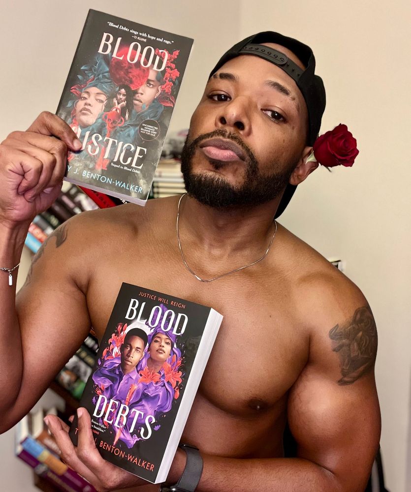 A photo of me shirtless holding copies of Blood Debts and Blood Justice. I have on a SnapBack and there’s a rose tucked behind one ear. 