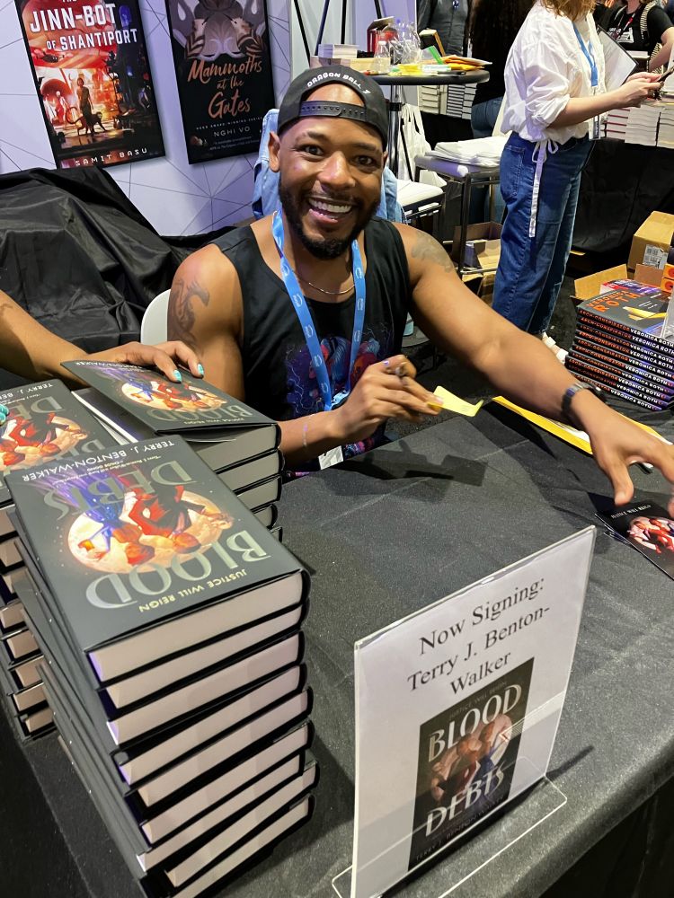 A photo of me at a signing table at SDCC signing copies of Blood Debts