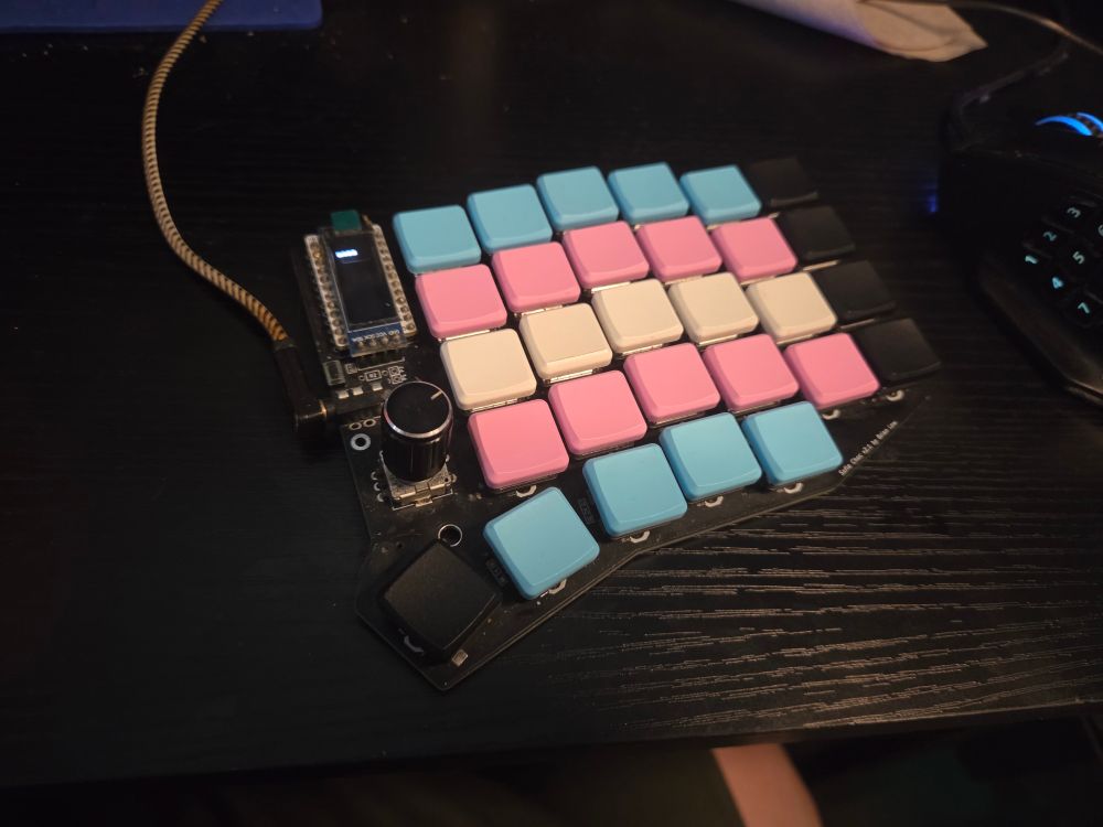 picture of the right half of a sofle v2 choc on a black desk
the oled says 'moss' but the fidelity isnt sufficient for readability in this image
the keycaps are blank and coloured to be a trans flag
the TRRS cable is yellowed from mustard incidents im messy ok
it is probably fairly dirty pleasE do not zoom in on my crUMBS

my keymap is also a little cursed