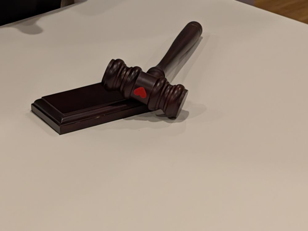 A dark brown wooden gavel resting on a block of the same material on a white table. The gavel has a red heart sticker on the top.