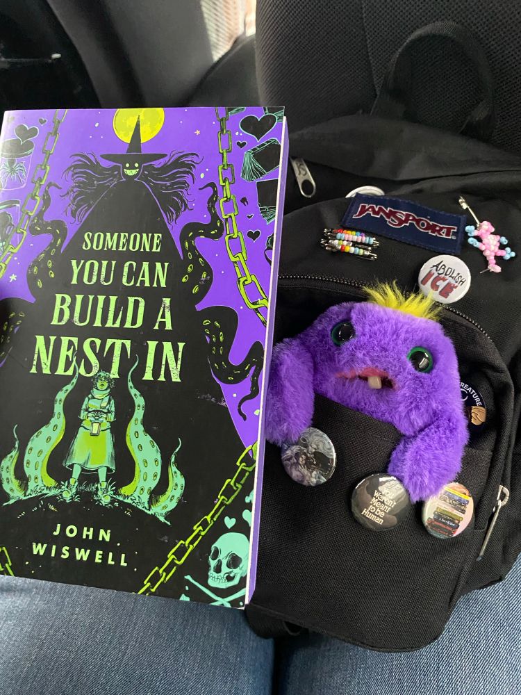 A photo of Someone You Can Build A Nest In by John Wiswell next to a black mini backpack with pins and a fuggler sticking out, they match in color