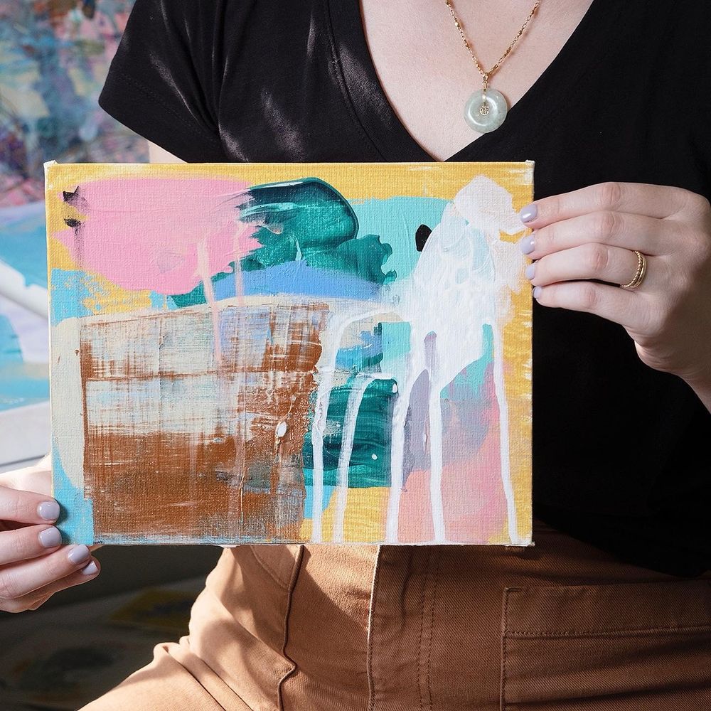 A close up photo where I am holding a medium sized abstract painting with golds, pinks, blues, greens and white paints. 