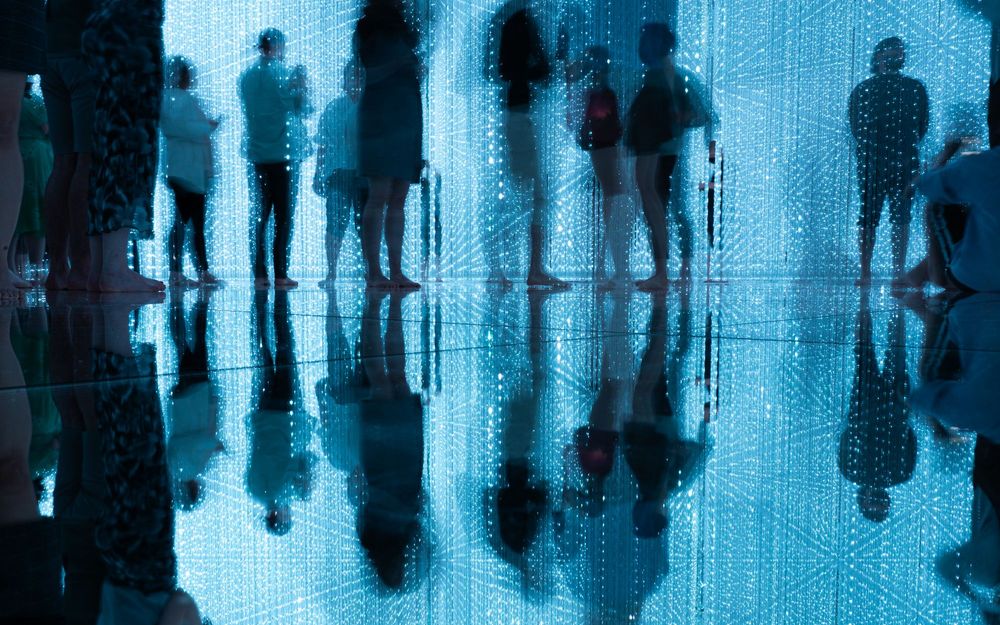 A slight long exposure photograph of people standing on a reflective floor at teamLab PLANETS in Tokyo. The people who have moved while the photograph was taken appear blurred, while the scene is backlit in a pale blue by hundreds of strips of LED lights hanging from the ceiling.