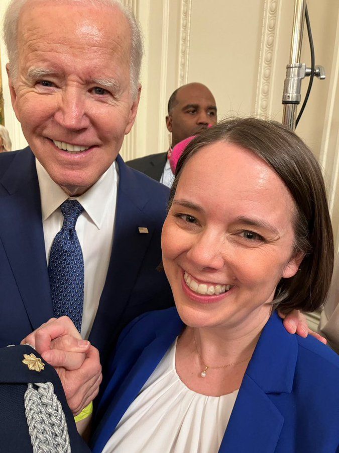Photo of President Biden holding hands with Maine Secretary of State Shenna Bellows, both wearing blue suits with white shirts. A Black man is visible behind them.