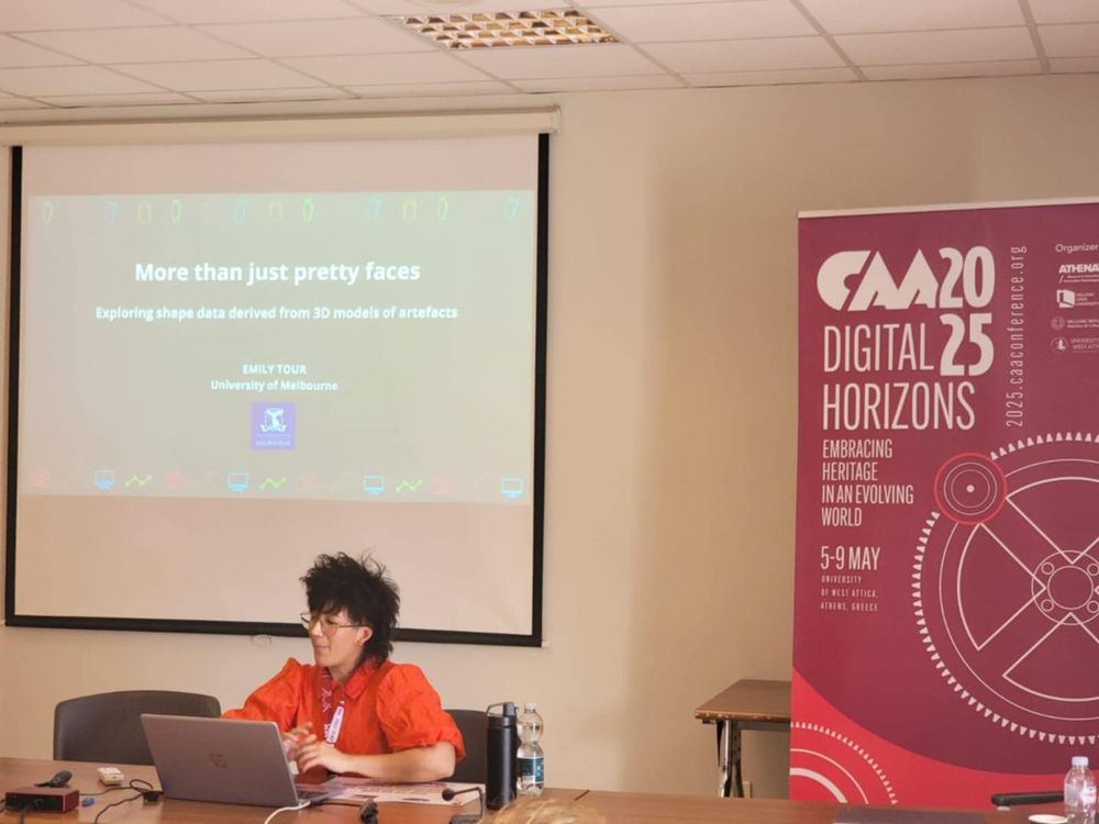 CAA Australasia member Emily Tour sitting at a table and presenting, with a slide presentation behind her reading 'More than just pretty faces: Exploring shape data derived from 3D models of artefacts.'