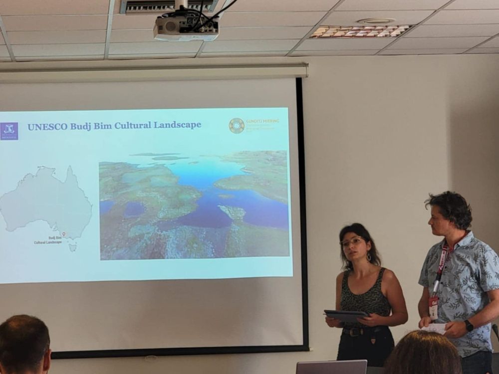 CAA Australasia members Aleks Michalewicz and Brian Armstrong standing in front of a slide presentation with images showing the UNESCO Budj Bim cultural landscape and its location within Australia.