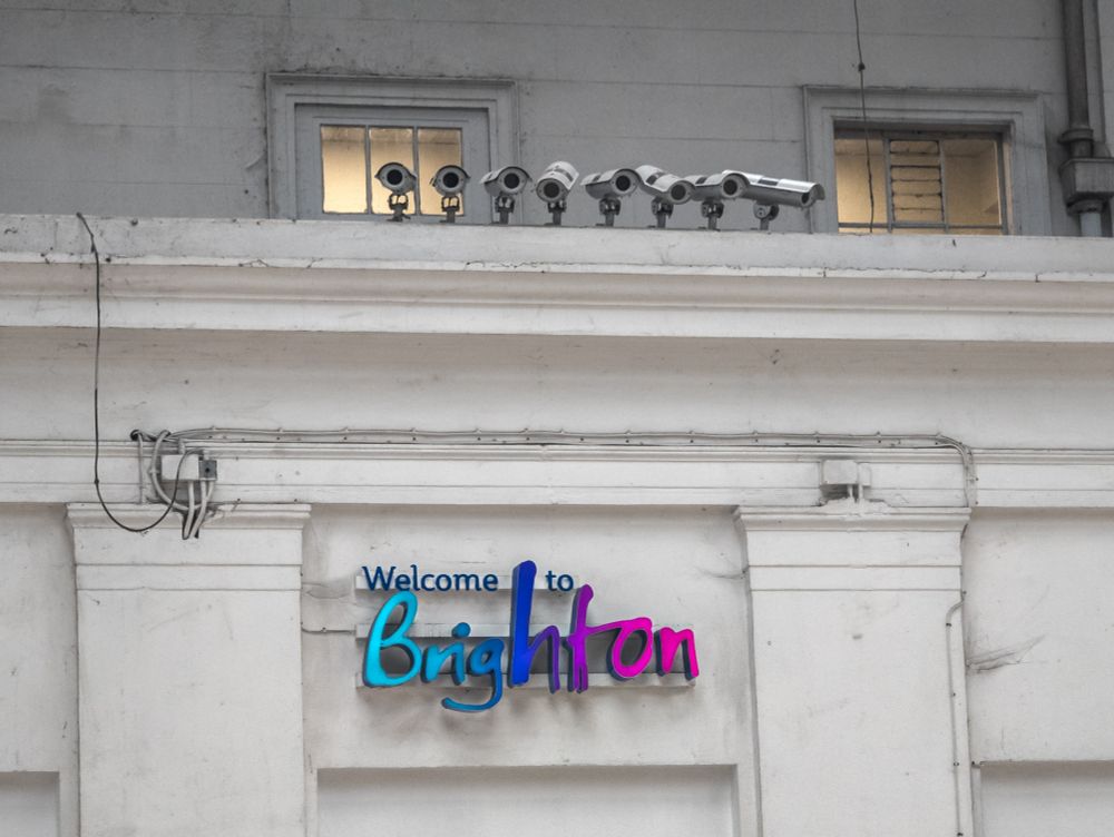 Above a 'Welcome to Brighton' sign lit in turquoise, blue and pink are a row of eight CCTV cameras pointing in different directions