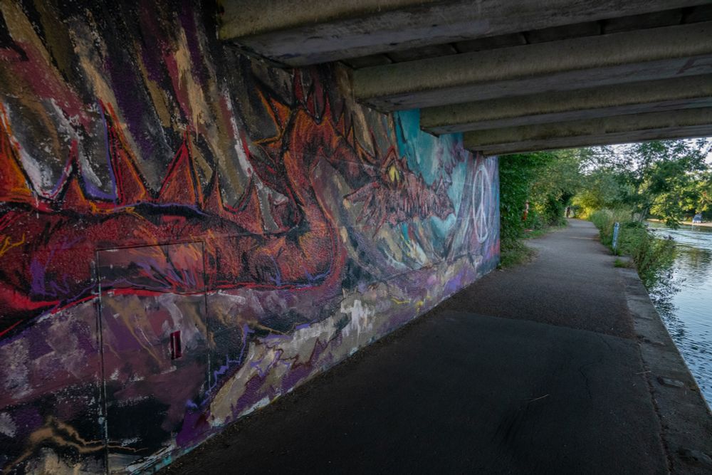 A painting of a red dragon, and a white peace sign is on the wall of a bridge by a river.
