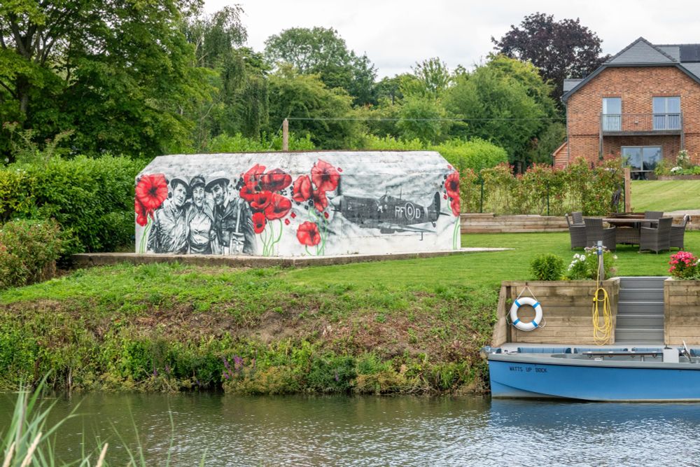 A concrete pillbox sits in a garden by a riverbank, it's painted white with an image of three soldiers arm in arm, red poppies, and a RAF plane on it. The garden has steps down to a boat, which is called "Watts Up Dock"