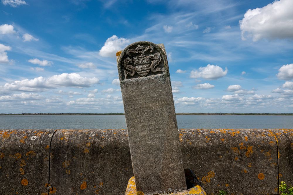 An ornate marker stone with a crest stands by a sea wall, in front of a wide river or estuary (the other side visible far away), with a blue sky in the background. The text reads:
"The eastern boundary of the juristdiction of the company of Watermen & Lightermen of the River Thames under the act of 22 & 23  VIC C 133
Erected 1861 Thomas White Master
Replaced 1982 Charles Peter Braithwaite Master."
Crest is two seahorses either side of a coat of arms.
