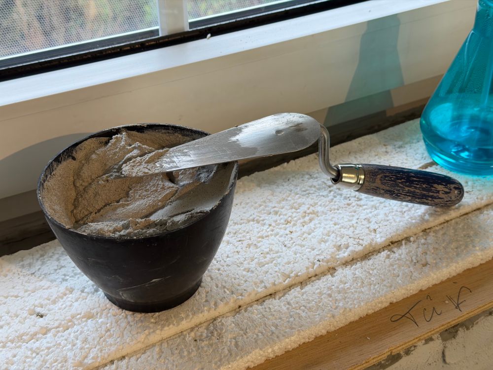 Auf dem Fenstersims steht ein Gipsbecher mit angerührtem Putz. Darauf liegt eine Kelle mit blau lackiertem Holzgriff. 