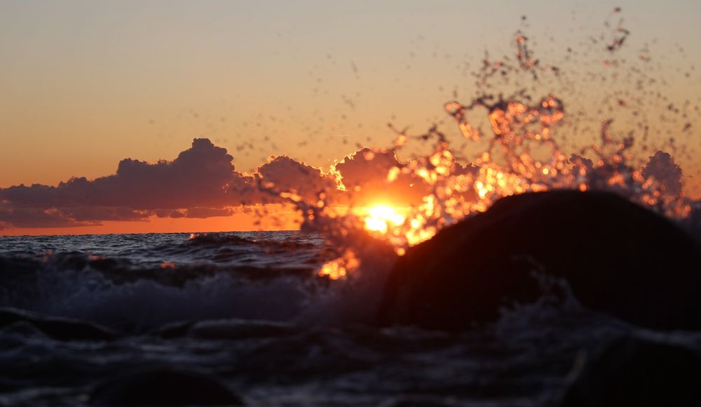 Eine Welle schwappt über einen Stein im Meer. Im Hintergrund Sonnenuntergang.