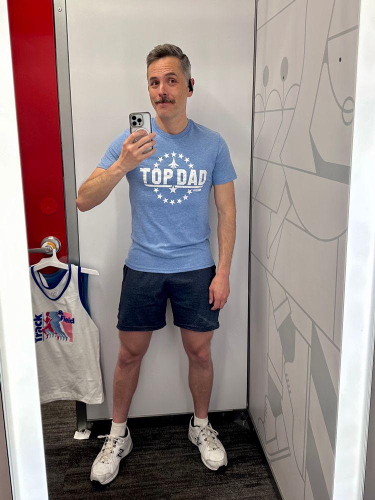 Mustached man wearing a blue T-shirt with the text “Top Dad” across the front in the style of Top Gun taking a selfie in a fitting room