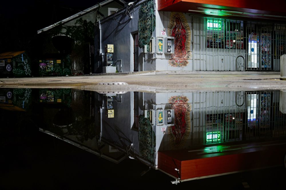 Corner view of gas station in the morning darkness with a lit sign and bright security light.  The top of frame is the station and the bottom of frame is the station’s reflection in a puddle. 