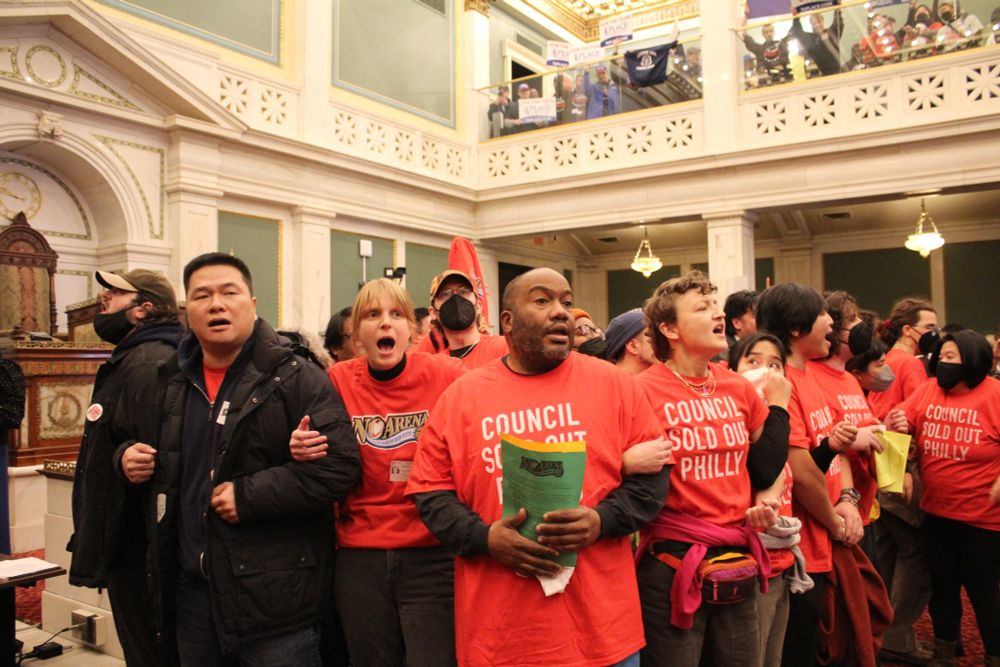 Multiracial group linking arms while taking over Philadelphia City Council chambers. They're wearing red t-shirts that say "Council Sold Out Philly"