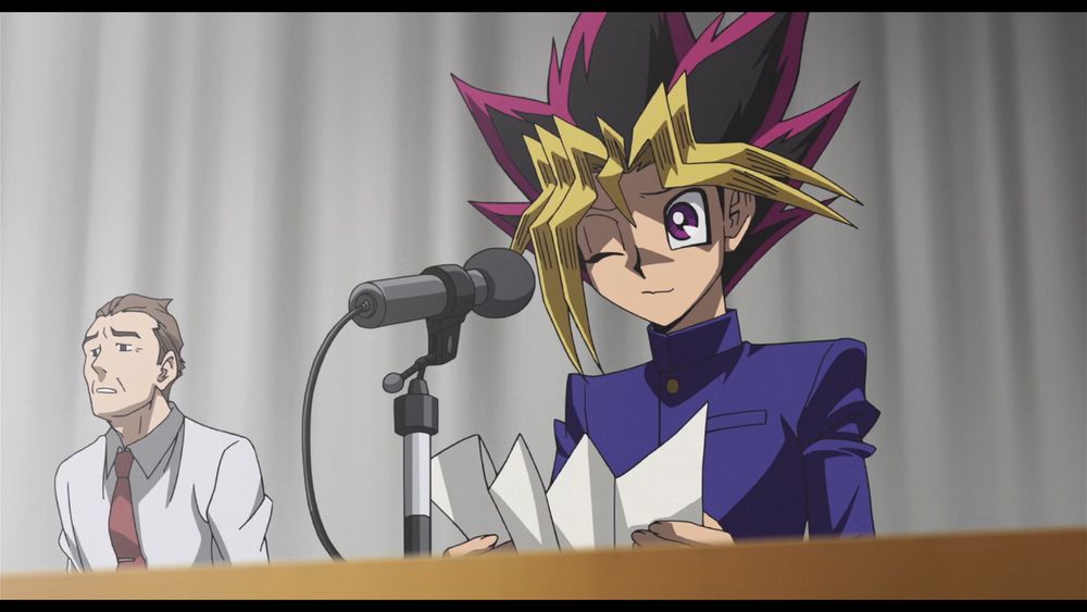 Screenshot from the movie Dark Side Of Dimensions. Yugi Muto is standing in front of a mic with a paper in his hand. He's winking/wincing.