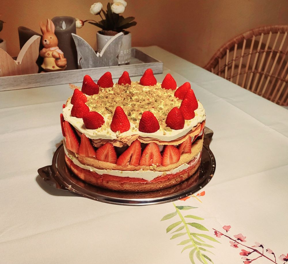 Eine Torte auf einem Esstisch. Die Torte besteht aus 3 hellen Böden mit einem Erdbeerfruchtspiegel, einer Schicht aus ganzen Erdbeeren, mehreren Schichten Pistazien-Mascarpone-Creme und Deko-Erdbeeren, sowie gehackten Pistazien als Deko