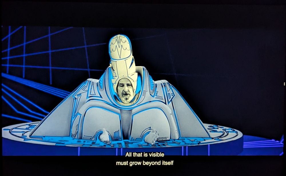 Man in a bizarre digital uniform from the movie Tron. He wears a headdress and large shoulder pads. He says "All that is visible must grow beyond itself."