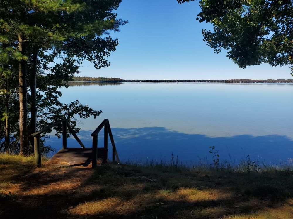 Midday view of a lake with light blue water and blue sky above, framed by pine and poplar trees.  A wooden staircase leads down to the lake.  The water closest to shore is shaded by the trees.  Beyond the shadow is a section of clear shallow water that shows the gold color sand on the bottom.  Across the lake is a line of trees, reflected in the still water.  