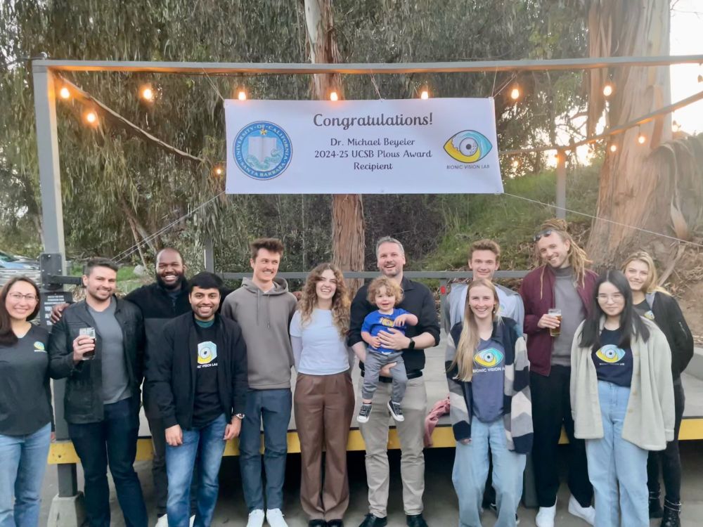 Michael Beyeler stands with members of the Bionic Vision Lab in front of a congratulatory banner celebrating his 2024–25 UCSB Plous Award. Everyone is smiling, with some holding drinks, and Michael is holding his young son. The group is gathered outdoors under string lights, with tall eucalyptus trees in the background.