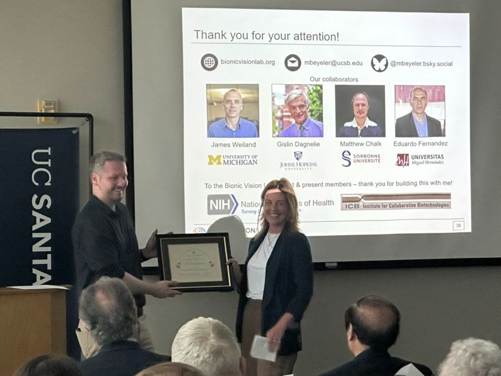 Michael Beyeler smiles while receiving the framed Plous Award at UC Santa Barbara. College of Letters & Science's Dean Shelly Gable presents the award in front of a slide thanking collaborators and funders, with photos of colleagues and logos from NIH and the Institute for Collaborative Biotechnologies. The audience watches the moment from their seats.