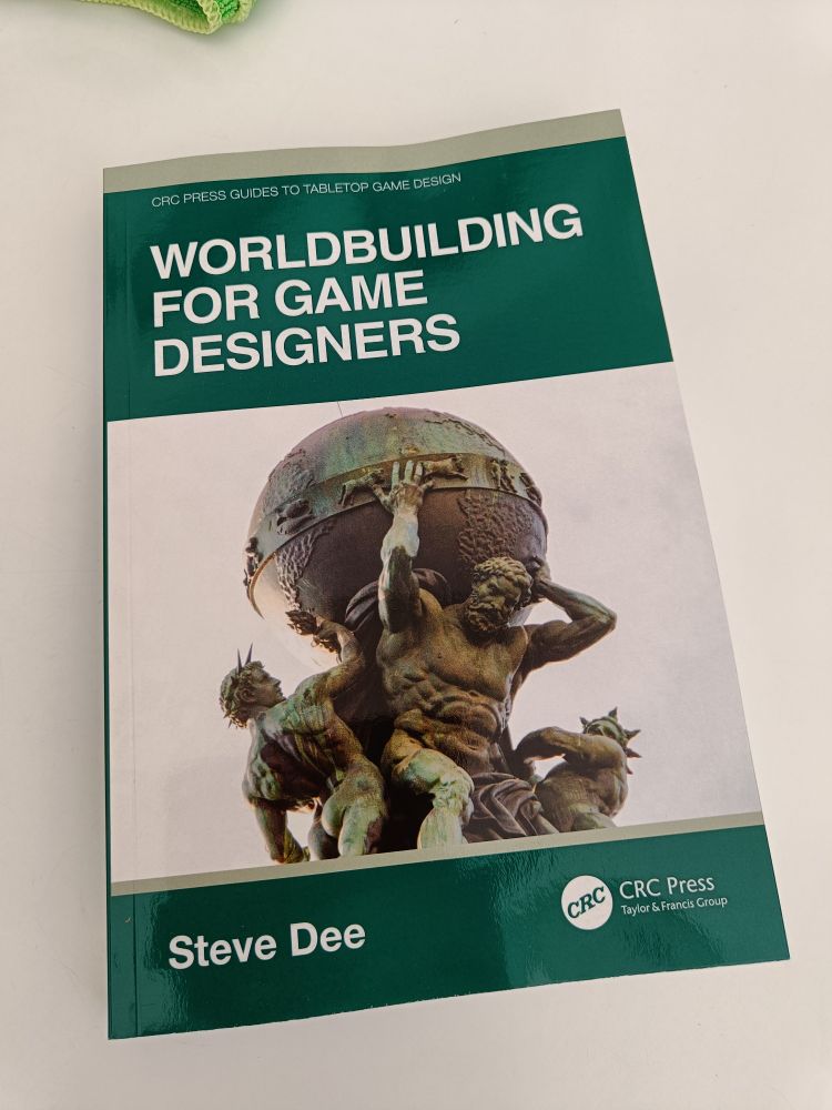 A chippy of the softback book Worldbuilding For Game Designers by Steve Dee. A green cover surrounds a photograph of a statue of Atlas holding up then globe of the Earth.