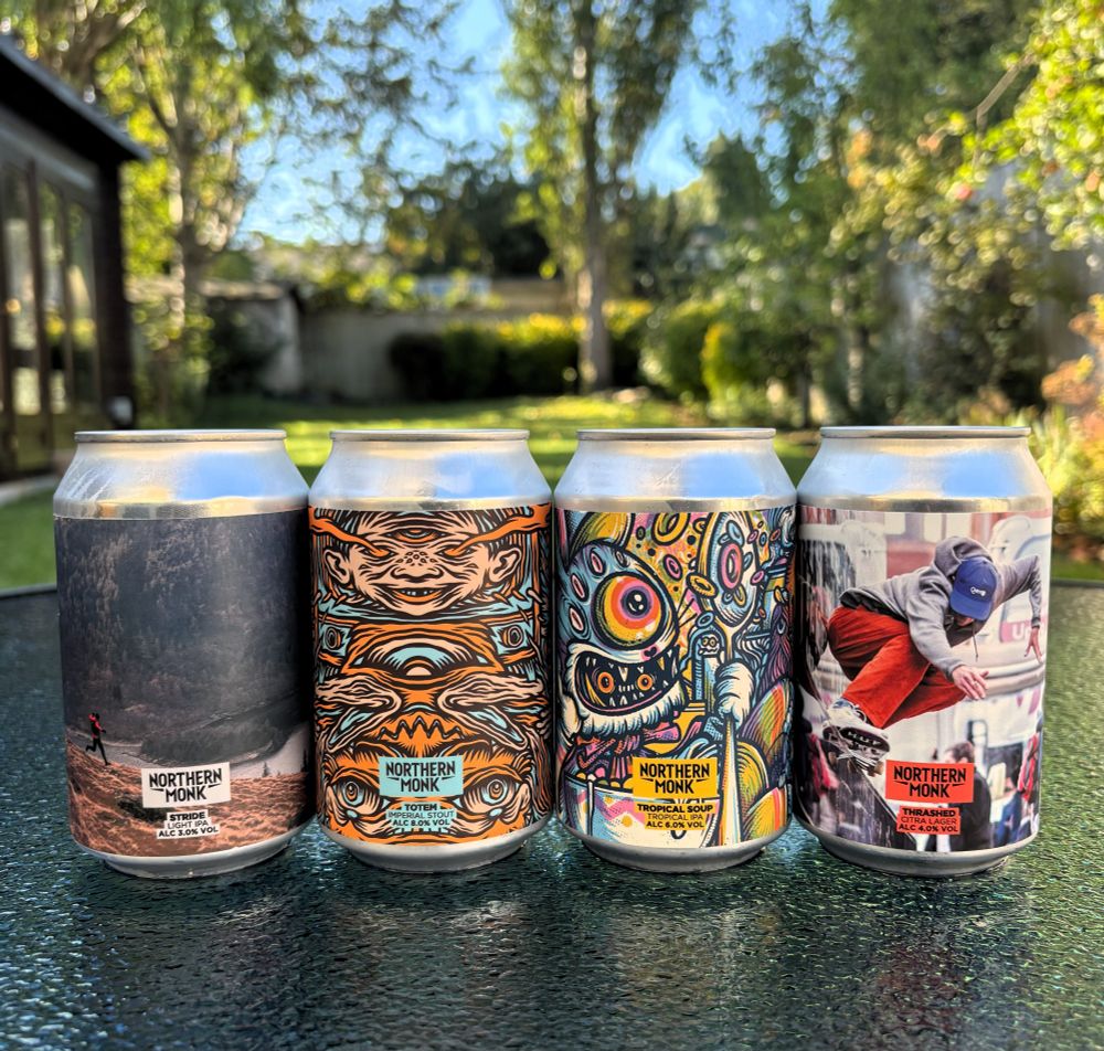 Four beer cans sitting on a rain-soaked glass table in a sunny garden. The beer cans have varying artwork. The beers are:

Northern Monk x Ricky Lightfoot - Stride Light IPA (3.0%)
Northern Monk x Tom J Newell - Totem Imperial Stout (8.0%)
Northern Monk x Drew Millard - Tropical Soup Tropical IPA (6.0%)
Northern Monk x Vague - Thrashed Citra Lager (4.0%)