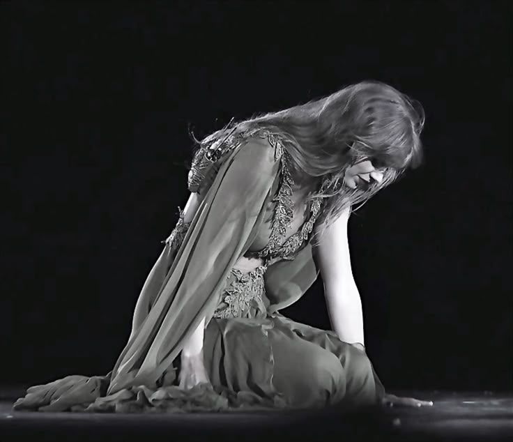 Taylor swift is kneeling on stage in her green folklore dress during her my tears ricochet Eras Tour performance. The photo is in black and white.
