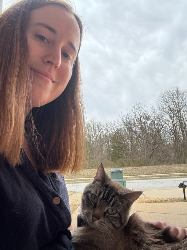 I am sitting outside my house with my neighbor’s tabby cat Tripp laying on my lap. I am wearing a blue jumpsuit and there are trees and our trash bin in the background.