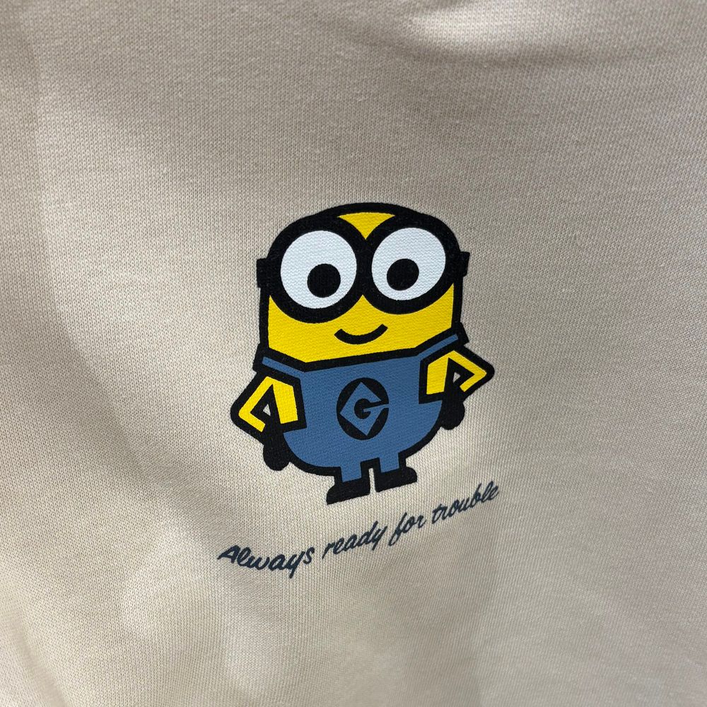 Auf einem Cremefarbenen Kleidungsstück ist ein gelber Minion in blauer Latzhose abgedruckt. Darunter steht in schwarzer Kursivschrift „Always ready for trouble“