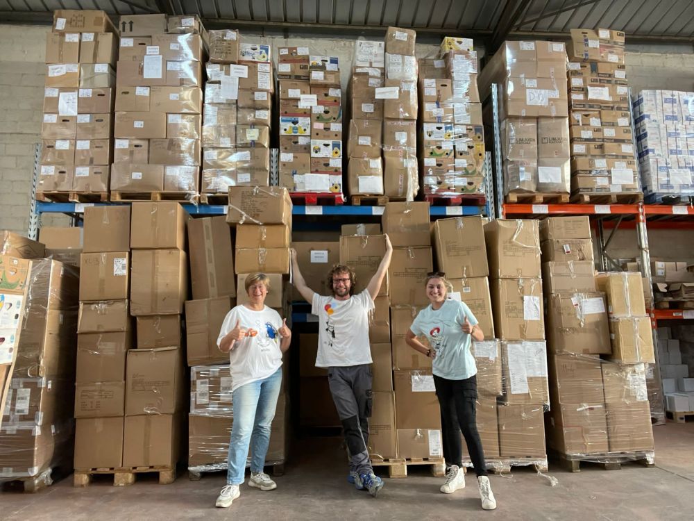 Three olunteers showing off neatly stacked pallets at their community aid warehouse.
