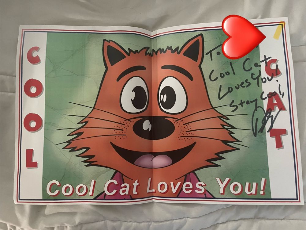 Poster of a cartoon orange cat. There is text on the poster that says “cool cat” and “cool cat loves you!” There is writing in black sharpie on the poster that says “To [redacted] cool cat loves you! Stay cool” with a signature underneath 