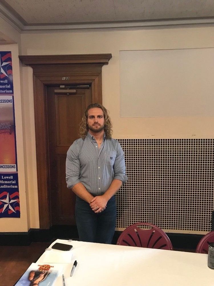 A picture of wrestler Hangman Adam Page standing politely with his hands grasped in front of him, wearing a button down shirt and jeans 