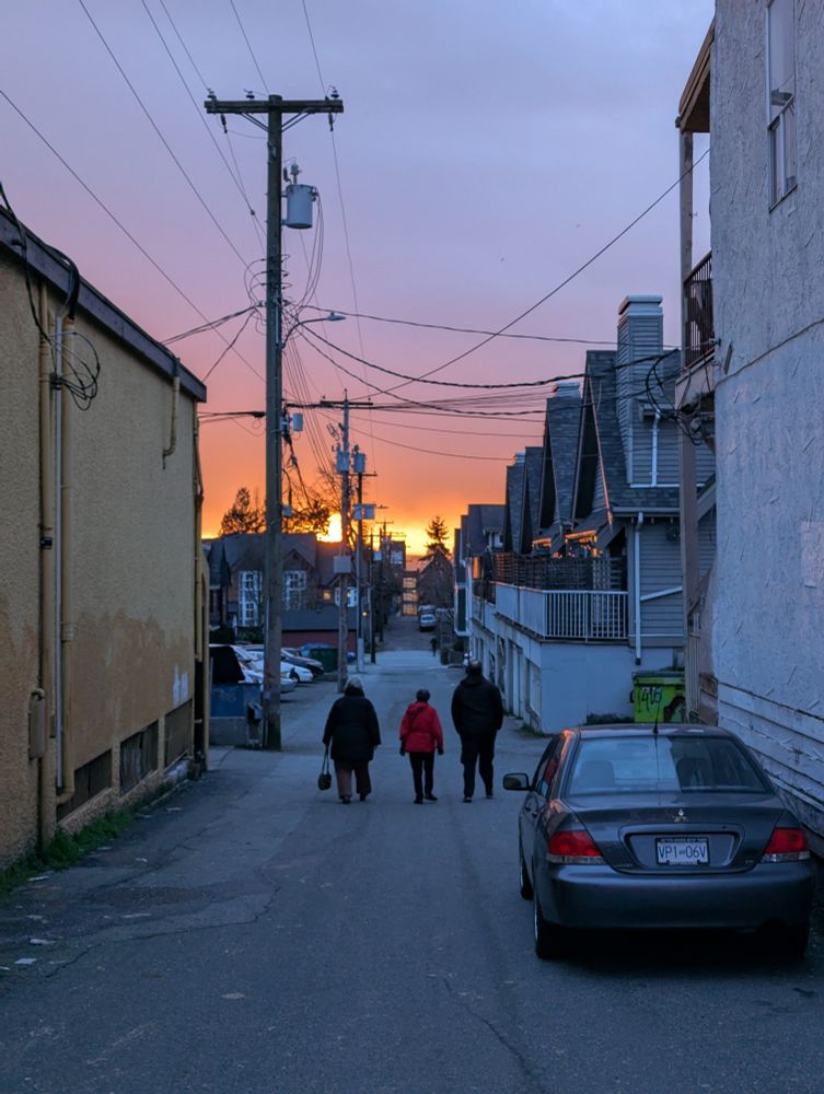 Three people walk down an alley towards a fiery sunset in East Vancouver.