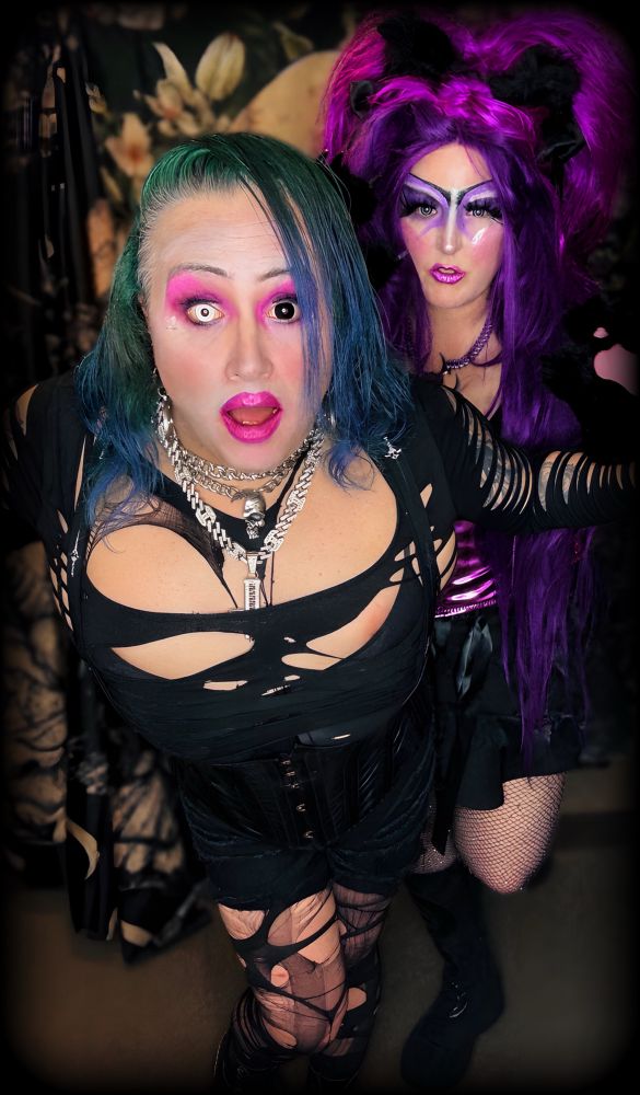 Two queers in bold, colorful makeup and dramatic, shredded black outfits pose together indoors, with one sporting teal hair and the other vibrant purple hair.