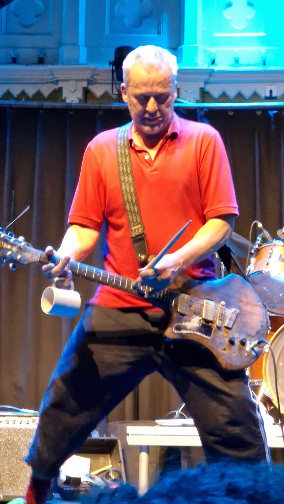 Terrie Ex bespeelt zijn gitaar met een theekop en een drumstok tijdens het optreden van The Ex op het podium van de grote zaal in Paradiso.