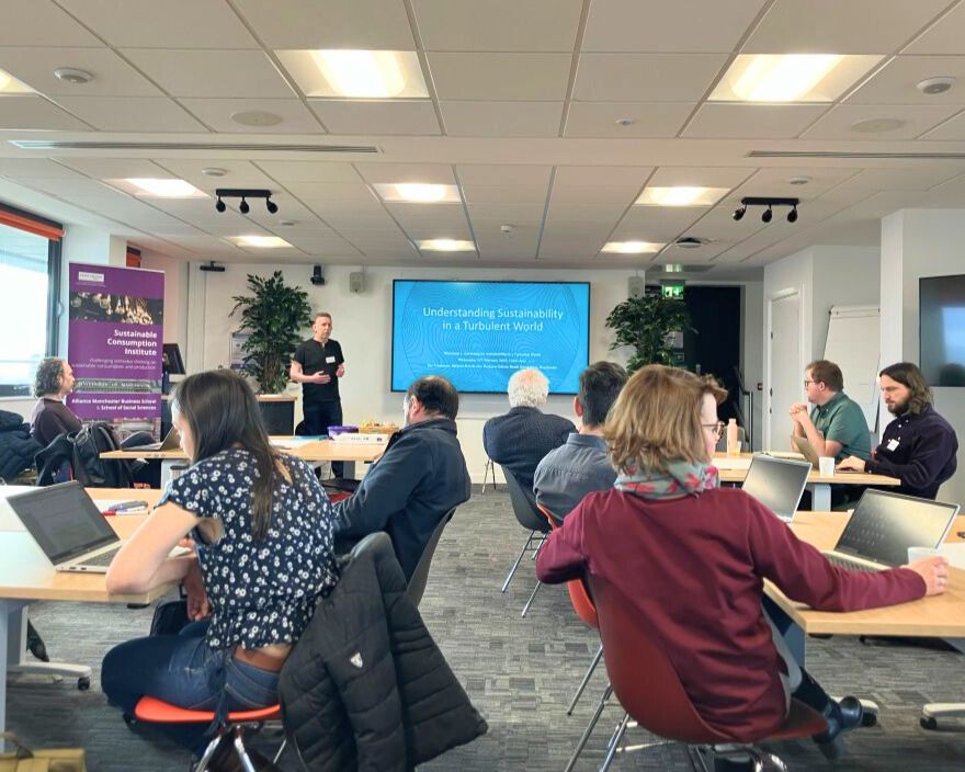 A group of people sat at different tables in a conference room with a blue presentation titled "Understanding Sustainability in a Turbulent World"