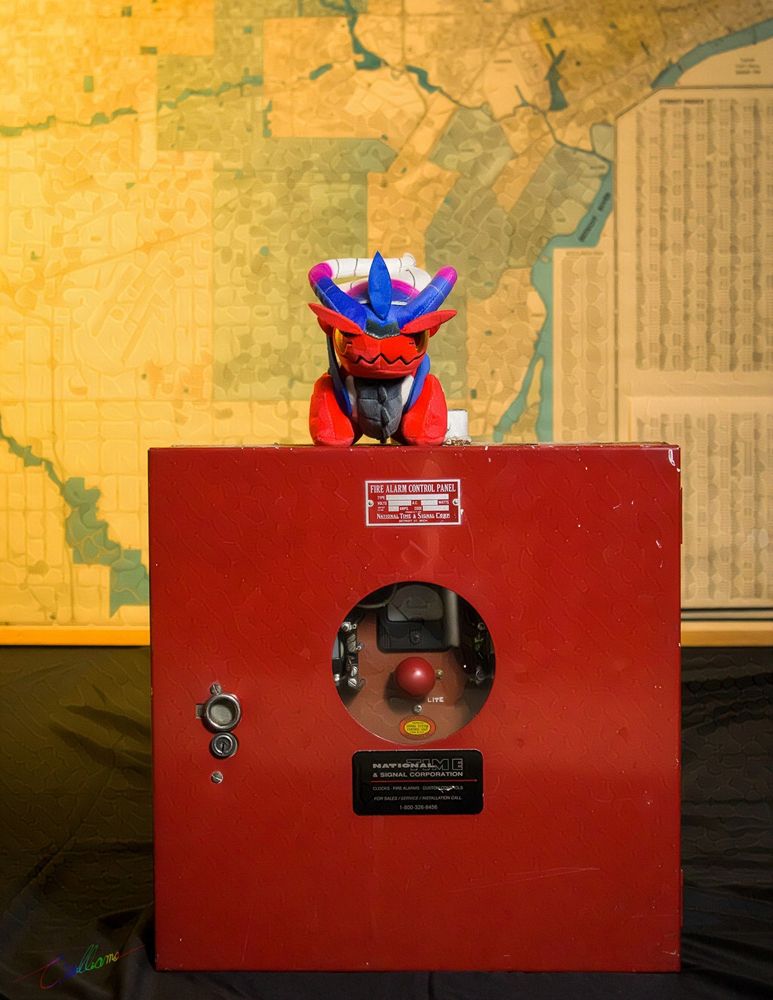Pokemon Koraidon plushie doll on top of a National Time & Signal Co 2124 fire alarm panel in front of a topographic map of Metro Detroit.