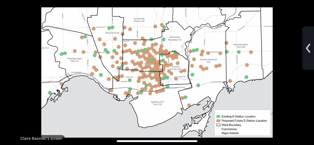 A geographical map of a city, primarily featuring a light gray and beige color scheme. The map outlines the city's boundaries, with distinct ward boundaries marked by black lines. Several major arteries and expressways are also highlighted. Numerous green and brown hexagonal markers are scattered across the map. These markers represent locations of existing and proposed future e-stations, with a legend providing clarification. The markers are concentrated in the central area of the map. The lower section of the map depicts a body of water, possibly a lake or ocean, and a coastal region. The overall style is illustrative and schematic, presenting a clear and informative visualization of the data. The perspective is an overhead view of the city. The color palette is muted, with the green and brown markers providing the only vibrant colors. The lighting is even, suggesting a straightforward representation.
