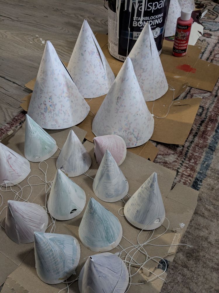 A bunch of white poorly painted birthday hats with a big can of primer and a little bottle of red acrylic paint in the background 