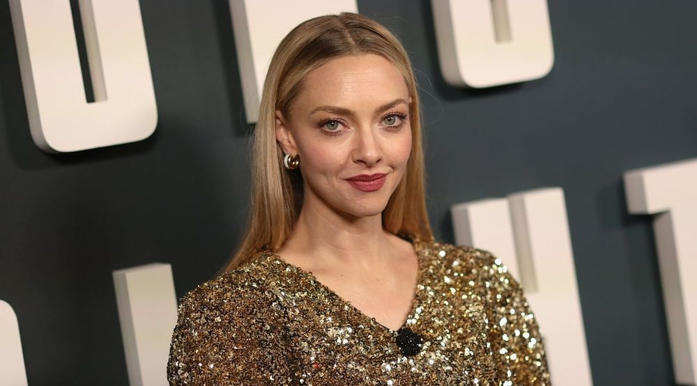Headshot of Amanda Seyfried at some event. She is wearing a glittering gold V neck. Her long blonde hair is styled straight and neatly tucked behind her ears