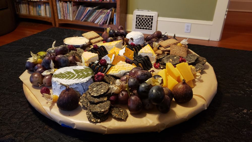 Round wooden board with several kinds of cheese, crackers, grapes, figs, and dries cherries on it. The board is on a table and the table is draped with a lacy black tablecloth. Only the top of the table with cloth is visible. In the background is a wall painted green, a bookcase,  and a couch.