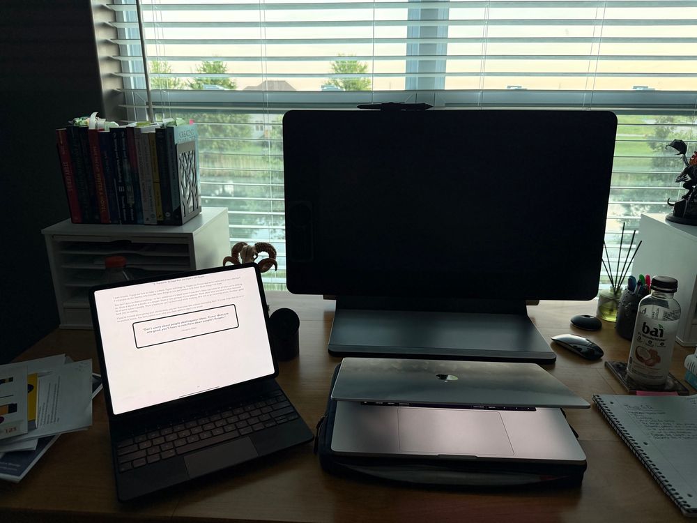 An iPad and a laptop on a desk with books and notebooks surrounding it and large black monitor in the center. Blinds for a window are open looking out onto a small pond. 
