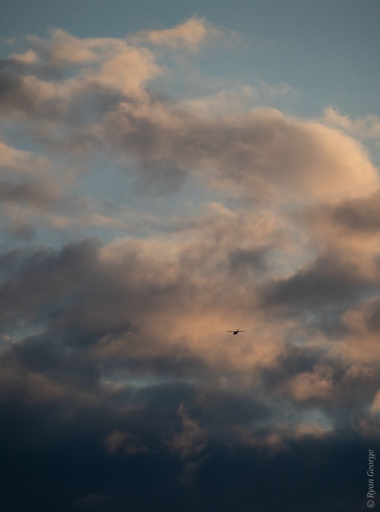 A photograph of multiple layers of clouds, illuminated by the setting sun. Near the middle of the clouds in a bright patch is the silhouette of a plane flying into the horizon. 