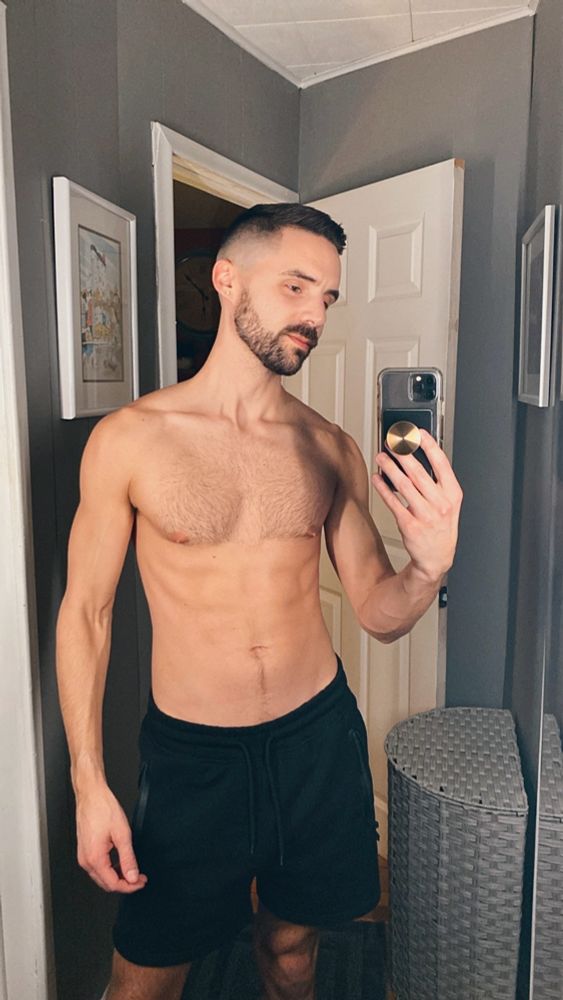 A shirtless man with a fresh haircut taking a selfie in a full-length mirror. 