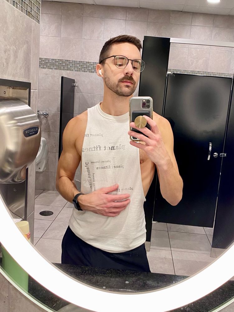 Man wearing a cut-off shirt taking a selfie in a bathroom mirror