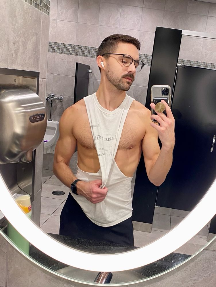 Man wearing a cut-off shirt, that’s pulled to the side to reveal his chest, taking a selfie in a bathroom mirror