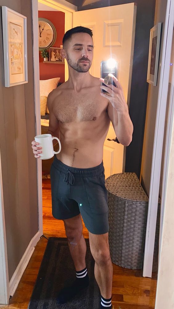 Shirtless man holding a coffee cup and taking a selfie in a hallway mirror