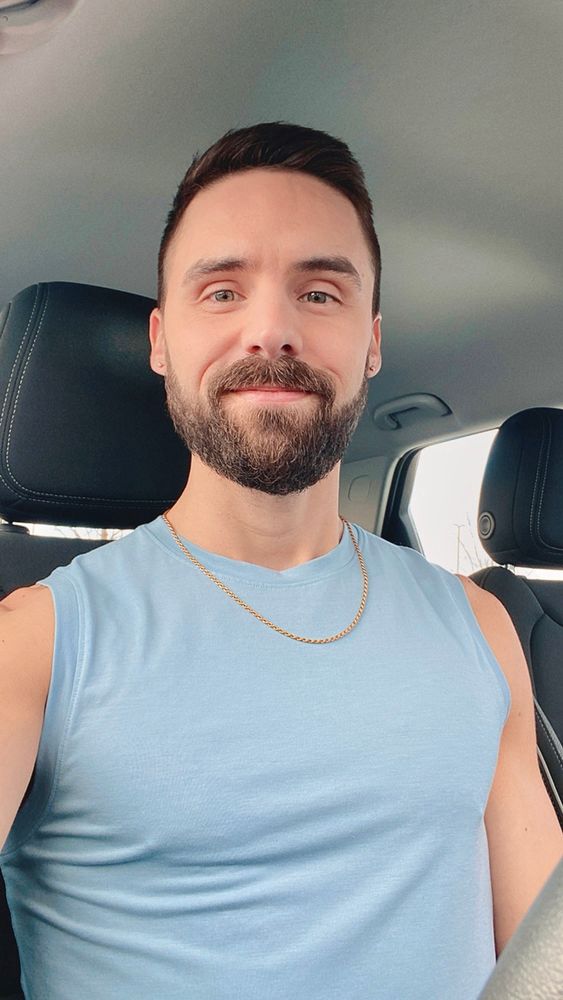 Selfie of a bearded man wearing a sleeveless shirt sitting in the front seat of a car.