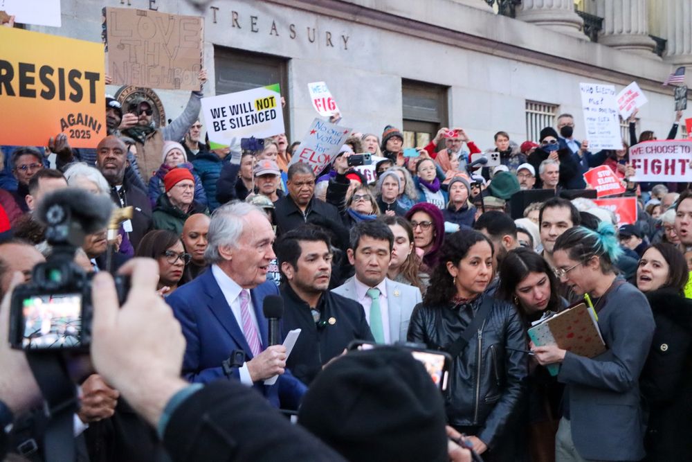 Senator Markey holds a microphone in front of a crowd holding resist and we will not be silenced and no one voted for Elon Musk signs